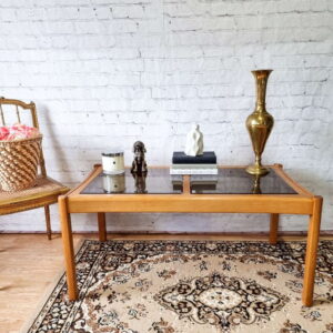 A room with a white brick wall features the Ercol Saville Ash Long Coffee Table with Smoked Glass Mid Century Vintage. Items on the table include a candle, a small sculpture, a stack of books, and a golden vase. A wicker chair with a cushion and a patterned rug are also visible.