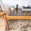 An Ercol Saville Ash Long Coffee Table with a smoked glass top is placed on a patterned rug. The table holds a candle, a small dog figurine, a white sculpture, and three stacked books. There is a rattan chair to the left and a white brick wall in the background.