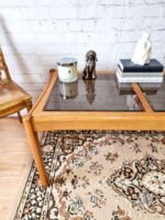 An Ercol Saville Ash Long Coffee Table with a smoked glass top is placed on a patterned rug. The table holds a candle, a small dog figurine, a white sculpture, and three stacked books. There is a rattan chair to the left and a white brick wall in the background.