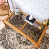 An Ercol Saville Ash Long Coffee Table with a smoked glass top is set on an ornate patterned rug. On the table, there are a few decor items including a candle, a small sculpture, and two stacked books. A brass lamp and a chair with a basket of pink flowers are nearby.