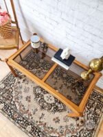 An Ercol Saville Ash Long Coffee Table with a smoked glass top is set on an ornate patterned rug. On the table, there are a few decor items including a candle, a small sculpture, and two stacked books. A brass lamp and a chair with a basket of pink flowers are nearby.