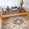 The Ercol Saville Ash Long Coffee Table with Smoked Glass Mid Century Vintage is placed on a patterned rug. It holds a candle, a decorative dog figurine, a stack of books with a white sculpture on top, and a tall brass vase. The background features a white brick wall.