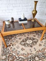 The Ercol Saville Ash Long Coffee Table with Smoked Glass Mid Century Vintage is placed on a patterned rug. It holds a candle, a decorative dog figurine, a stack of books with a white sculpture on top, and a tall brass vase. The background features a white brick wall.