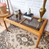 An Ercol Saville Ash Long Coffee Table with Smoked Glass Mid Century Vintage is set in a room with a white brick wall. On the table are books, a brass vase, a candle, a decorative statue, and a small figurine of a dog. To the left is a chair with a basket of pink roses on it. The floor is covered by an ornate rug.