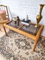 An Ercol Saville Ash Long Coffee Table with Smoked Glass Mid Century Vintage is set in a room with a white brick wall. On the table are books, a brass vase, a candle, a decorative statue, and a small figurine of a dog. To the left is a chair with a basket of pink roses on it. The floor is covered by an ornate rug.