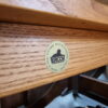 Close-up of an **Ercol Saville Ash Long Coffee Table with Smoked Glass Mid Century Vintage** featuring a circular gold sticker that reads "Ercol" and "Made in High Wycombe, Buckinghamshire" around the edge. The wood grain is visible, showcasing a medium brown finish.