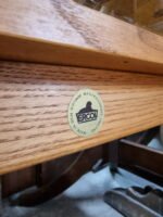 Close-up of an **Ercol Saville Ash Long Coffee Table with Smoked Glass Mid Century Vintage** featuring a circular gold sticker that reads "Ercol" and "Made in High Wycombe, Buckinghamshire" around the edge. The wood grain is visible, showcasing a medium brown finish.