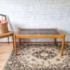 An Ercol Saville Ash Long Coffee Table with Smoked Glass Mid Century Vintage is placed on a patterned rug featuring beige and brown designs. To the left, a woven basket filled with pink flowers rests on a wooden chair with a cane seat. The background showcases a white brick wall.