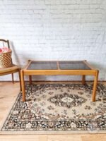 An Ercol Saville Ash Long Coffee Table with Smoked Glass Mid Century Vintage is placed on a patterned rug featuring beige and brown designs. To the left, a woven basket filled with pink flowers rests on a wooden chair with a cane seat. The background showcases a white brick wall.