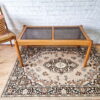 The Ercol Saville Ash Long Coffee Table with Smoked Glass Mid Century Vintage sits on an ornate, patterned rug. The background features a white brick wall and part of a wicker chair is visible on the left side of the image.