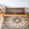 An Ercol Saville ash long coffee table with a smoked glass top is placed on an ornate, patterned rug. The table has a simple rectangular design and is positioned against a white painted brick wall. A wooden chair with a woven seat and backrest is partially visible on the left.