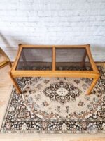 An Ercol Saville ash long coffee table with a smoked glass top is placed on an ornate, patterned rug. The table has a simple rectangular design and is positioned against a white painted brick wall. A wooden chair with a woven seat and backrest is partially visible on the left.