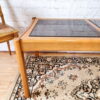 An Ercol Saville Ash Long Coffee Table with a smoked glass top is situated on a patterned rug. Beside it is a wicker chair with a wooden frame. The background features a white brick wall, creating a bright and airy ambiance. Part of the chair and the table legs are visible in the forefront.