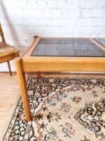 An Ercol Saville Ash Long Coffee Table with a smoked glass top is situated on a patterned rug. Beside it is a wicker chair with a wooden frame. The background features a white brick wall, creating a bright and airy ambiance. Part of the chair and the table legs are visible in the forefront.