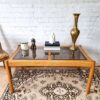 An Ercol Saville Ash Long Coffee Table with a smoked glass top sits on an ornate rug. On the table are a candle, a small statuette, a stack of books, a white sculpture, and a tall brass vase. Behind the table is a white brick wall.