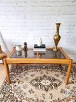 An Ercol Saville Ash Long Coffee Table with a smoked glass top sits on an ornate rug. On the table are a candle, a small statuette, a stack of books, a white sculpture, and a tall brass vase. Behind the table is a white brick wall.