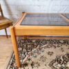 An Ercol Saville Ash Long Coffee Table with a smoked glass top sits gracefully on a detailed, patterned rug. The brick wall and wooden chair with a woven seat in the background contribute to the rustic ambiance. The rug showcases an intricate design featuring shades of brown, beige, and black.