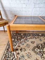 An Ercol Saville Ash Long Coffee Table with a smoked glass top sits gracefully on a detailed, patterned rug. The brick wall and wooden chair with a woven seat in the background contribute to the rustic ambiance. The rug showcases an intricate design featuring shades of brown, beige, and black.