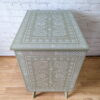 A light green Faux Bone Inlay Stencil Hand-Painted Vintage Chest of Drawers adorned with intricate white floral and geometric patterns on the top and front. The chest stands gracefully on four legs, placed on a wooden floor against a white brick wall, with the "Lily & Design" logo visible in the bottom right corner.