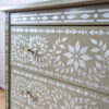 A Faux Bone Inlay Stencil Hand-Painted Vintage Chest of Drawers in green, adorned with intricate white floral and geometric patterns, featuring three drawers with small, round metallic handles. The background showcases a white brick wall, and part of a wooden floor is visible at the bottom.