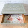 A Faux Bone Inlay Stencil Hand-Painted Vintage Chest of Drawers, featuring a square green design with a patterned white floral motif on the top and sides. This chest boasts an open drawer adorned with decorative paper showcasing birds and floral designs. The chest is displayed against a white brick wall on a wooden floor.