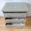 A small Faux Bone Inlay Stencil Hand-Painted Vintage Chest of Drawers stands against a white brick wall. The chest features intricate white stencil designs on the top and drawers. Two lower drawers are pulled open, revealing a floral-patterned lining inside. The wood floor complements the chest.