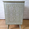 A Faux Bone Inlay Stencil Hand-Painted Vintage Chest of Drawers, painted in green and adorned with intricate white stencil patterns featuring floral and geometric designs, stands on tapered legs. This elegant chest is set against a wooden floor with a white brick wall in the background.