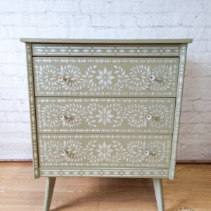 The Faux Bone Inlay Stencil Hand-Painted Vintage Chest of Drawers is a small, green wooden chest with three drawers, each adorned with intricate white floral patterns. The chest stands on four tapered legs and is showcased against a white brick wall background on a hardwood floor.