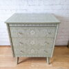 A small Faux Bone Inlay Stencil Hand-Painted Vintage Chest of Drawers in green, featuring three drawers adorned with intricate white floral and geometric patterns. The dresser stands on tapered legs against a white brick wall, with a textured wood floor beneath.