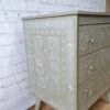 A Faux Bone Inlay Stencil Hand-Painted Vintage Chest of Drawers, showcasing intricate white floral and geometric designs against a light green backdrop, stands gracefully against a white brick wall. The chest features three drawers, each adorned with a small round metallic knob. The wooden floor beneath enhances the warmth of the setup.