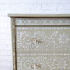 The Faux Bone Inlay Stencil Hand-Painted Vintage Chest of Drawers is a light green dresser featuring three drawers, each adorned with intricate white floral patterns and round white knobs. Set against a white brick wall, the muted color and detailed design of the furniture create a striking contrast.