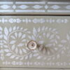 A close-up image of a drawer from the Faux Bone Inlay Stencil Hand-Painted Vintage Chest of Drawers, featuring intricate white floral and leaf patterns on a beige background. The drawer has a single, circular, bronze-colored knob in the center. A small logo is visible in the bottom right corner of the image.