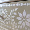 Close-up of the Faux Bone Inlay Stencil Hand-Painted Vintage Chest of Drawers, showcasing an intricate white floral and geometric pattern on a pale olive green background. The design features leaves, flowers, and circular motifs, giving a detailed, decorative appearance to the surface.