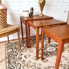 The Ercol Saville Ash Long Coffee Table with Smoked Glass Mid Century Vintage is arranged on a patterned rug. A brass lamp, a white candle, and a small bronze figurine are displayed on the table. An ornate wicker basket is placed on a decorative chair to the left. The background features a white brick wall.