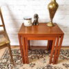 A beautifully arranged Ercol Saville Ash long coffee table with smoked glass rests on a patterned rug, holding a brass lamp, a scented candle, and a small dog figurine. To the left is the side view of a gold chair, and in the background stands a white brick wall.