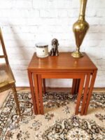 A beautifully arranged Ercol Saville Ash long coffee table with smoked glass rests on a patterned rug, holding a brass lamp, a scented candle, and a small dog figurine. To the left is the side view of a gold chair, and in the background stands a white brick wall.