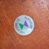 A silver circular sticker with green and purple overlapping triangles in the center. The text around the edge reads "Furniture by McIntosh." The sticker is attached to an Ercol Saville Ash Long Coffee Table with Smoked Glass Mid Century Vintage, featuring a rich, brown finish.