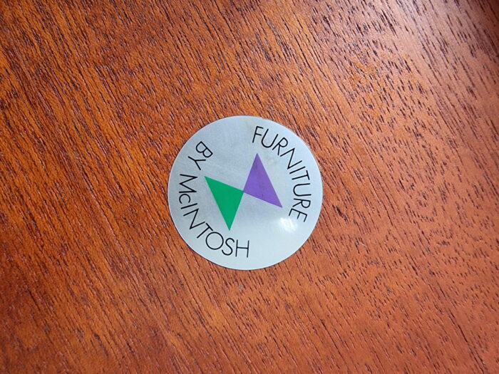 A silver circular sticker with green and purple overlapping triangles in the center. The text around the edge reads "Furniture by McIntosh." The sticker is attached to an Ercol Saville Ash Long Coffee Table with Smoked Glass Mid Century Vintage, featuring a rich, brown finish.