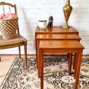 An Ercol Saville Ash Long Coffee Table with Smoked Glass Mid Century Vintage features prominently against a white brick wall, topped with a brass vase, a candle, and a small statuette. To the left of this setup is a wooden chair with a woven seat and a basket of pink roses, all elegantly placed on an ornate patterned rug.