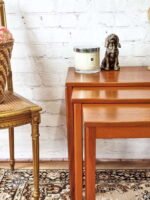 A white brick wall serves as the backdrop for an Ercol Saville Ash Long Coffee Table with Smoked Glass Mid Century Vintage. On the table, there's a candle and a dog-shaped bronze figurine. To the left, a wicker basket with pink flowers sits on a vintage-style chair with a gold frame. A patterned rug is on the floor.