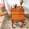 The Ercol Saville Ash Long Coffee Table with Smoked Glass Mid Century Vintage on a patterned rug, accompanied by a brass lamp, a ceramic container with a lid, and a small dog figurine. To the left, a wicker basket filled with pink roses sits on a wooden chair against a white brick wall.