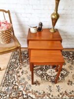 The Ercol Saville Ash Long Coffee Table with Smoked Glass Mid Century Vintage on a patterned rug, accompanied by a brass lamp, a ceramic container with a lid, and a small dog figurine. To the left, a wicker basket filled with pink roses sits on a wooden chair against a white brick wall.