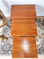 An Ercol Saville Ash Long Coffee Table with Smoked Glass sits elegantly on a decorative rug, with a white brick wall serving as the backdrop. A portion of a brass lamp and a framed picture are partially visible to the left, adding to the vintage mid-century ambiance.