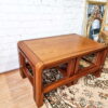 A Mid Century Teak Nest of Tables, featuring a coffee table and two side tables, sits on a patterned beige rug. Next to it, leaning against a white brick wall, are ornate gold-framed mirrors and a chair with similar wood design.