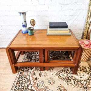 A set of Mid Century Teak Nest of Tables, with a coffee table and two side tables, stands on a patterned rug in front of a white brick wall. The coffee table holds a vase, two small decorative items, a stack of books, and additional decor pieces nearby. The tables feature a minimalist design with open lower shelves.