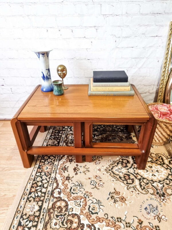 A set of Mid Century Teak Nest of Tables, with a coffee table and two side tables, stands on a patterned rug in front of a white brick wall. The coffee table holds a vase, two small decorative items, a stack of books, and additional decor pieces nearby. The tables feature a minimalist design with open lower shelves.