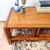 The Mid Century Teak Nest of Tables - Coffee Table and Two Side Tables display a striking blue and white vase, a petite decorative sculpture, a green candle holder, and several stacked books. The tables rest on a beige patterned rug set against a white brick wall background.