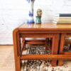 A Mid Century Teak Nest of Tables - Coffee Table and Two Side Tables set with a glossy finish is placed against a white brick wall. On the tables are books, a blue and white porcelain vase, a small green statue, and another small gold sculpture. A patterned rug is on the floor under the tables.