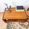 The Mid Century Teak Nest of Tables, consisting of a coffee table and two side tables, is decorated with a blue and white vase, a small brass globe candle holder, a green ceramic cup, and a stack of books. The set is arranged on a patterned carpet against a white brick wall. Pink flowers rest on the floor beside the tables.