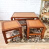 A set of three Mid Century Teak Nest of Tables, featuring a clean and minimalist design, are placed on a patterned area rug. The two smaller square side tables are nested under the larger rectangular coffee table. A gilded ornate chair and a large decorative mirror adorn the background against a white brick wall.